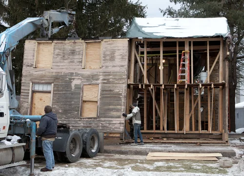 Demolition of the original 1857 house in winter, with workers removing wood siding next to a flatbed truck