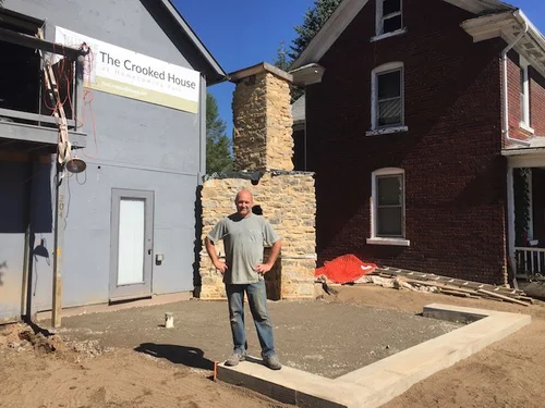 Benjamin Fehl standing in front of the stone chimney during construction, with The Crooked House banner above