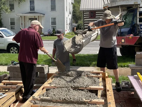 Three volunteers pouring concrete from a skid-steer bucket into the sidewalk forms
