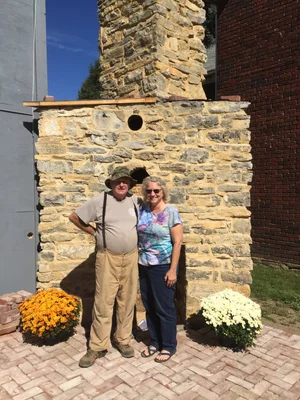 Two visitors posing in front of the completed stone chimney, with potted mums at its base