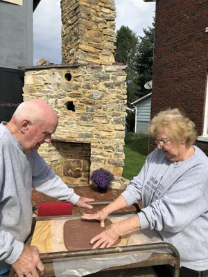 Two volunteers shaping clay tiles at an outdoor workshop in front of the stone chimney